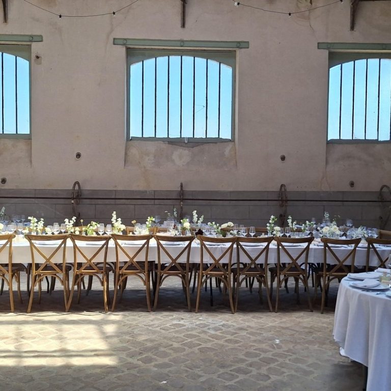 Grande salle avec tables dressées pour un banquet, fenêtres laissant entrer la lumière.