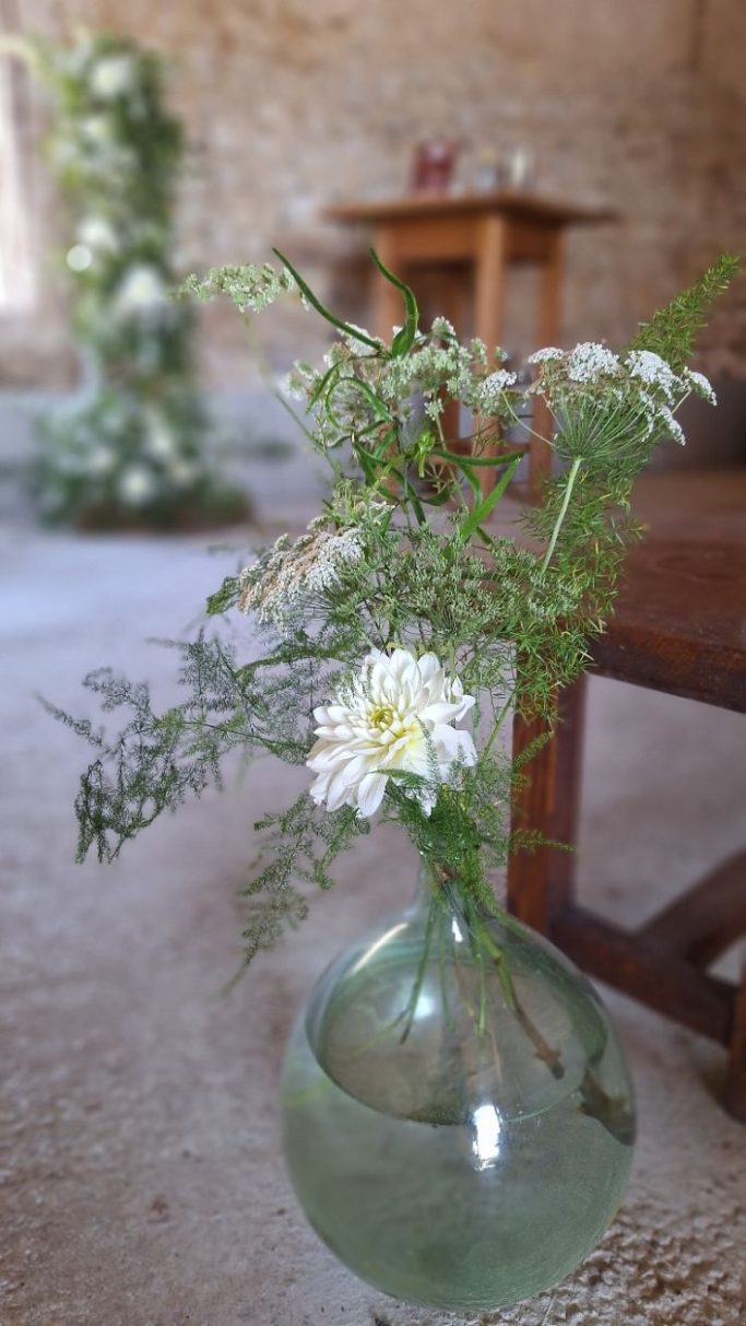 Vase en verre avec des fleurs blanches et des feuillages verts dans un intérieur rustique.