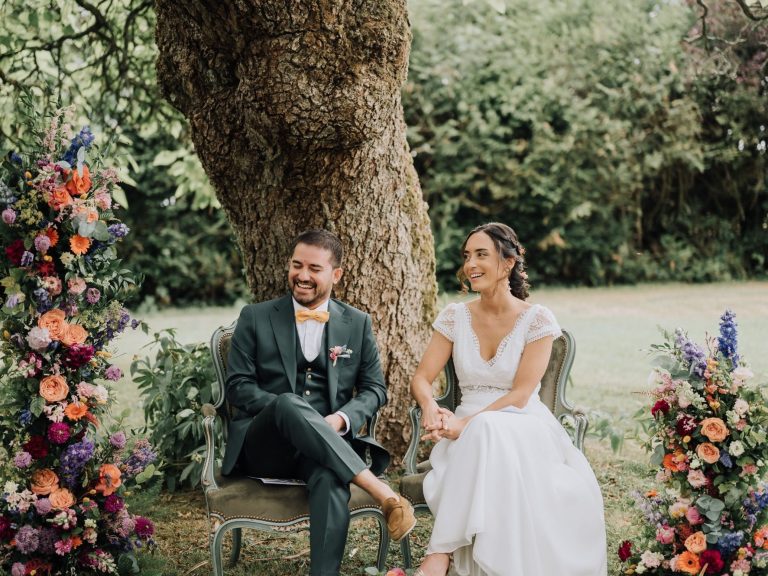 Mariage en plein air avec un couple assis sous un arbre, entouré de fleurs colorées.