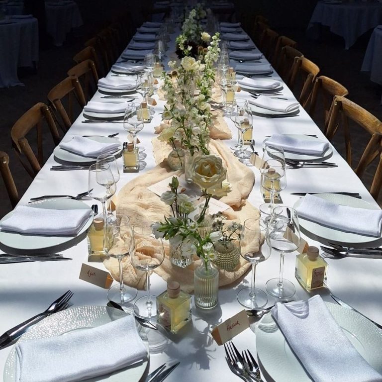 Longue table dressée avec nappes blanches, fleurs et couverts élégants.