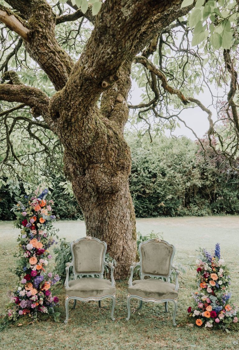 Deux chaises en velours au pied d'un arbre, entourées de fleurs colorées.