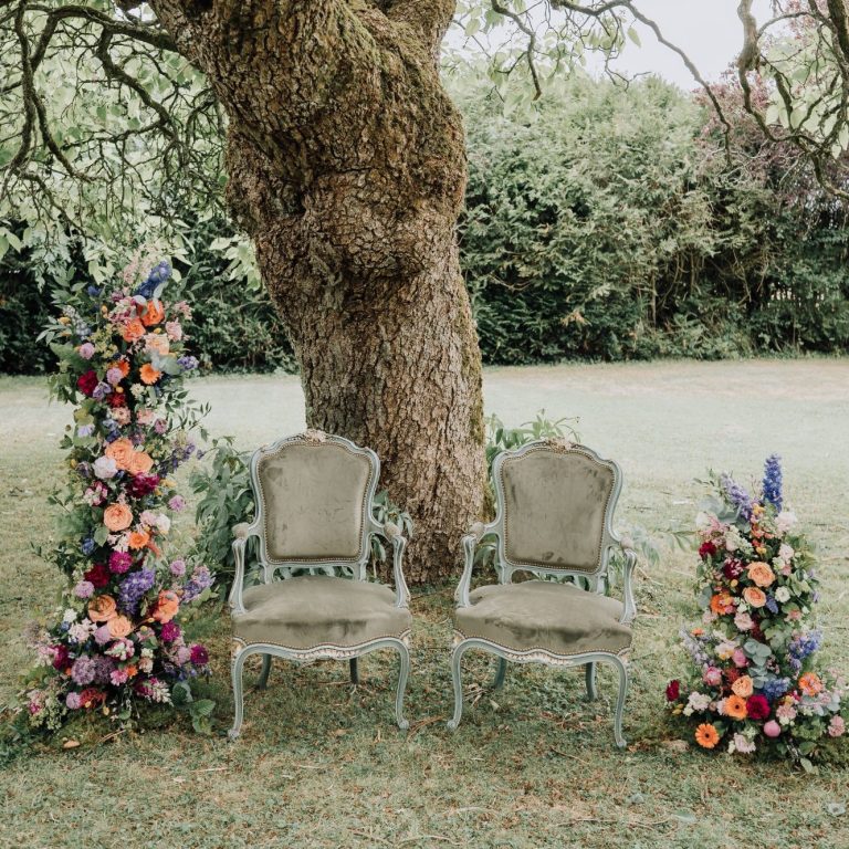 Deux chaises vintage vertes encadrées de bouquets floraux colorés sous un arbre.