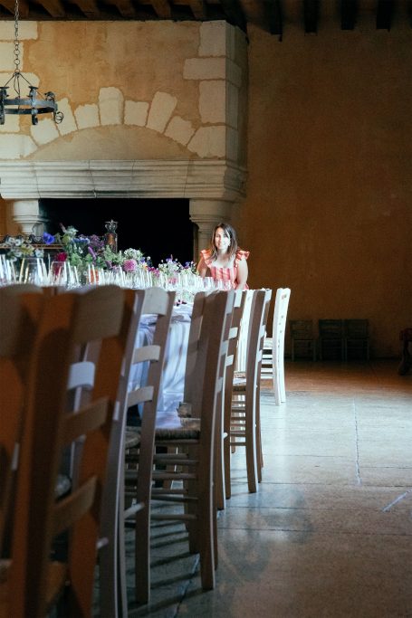 Une grande table dressée avec des chaises vides, une femme assise au fond.