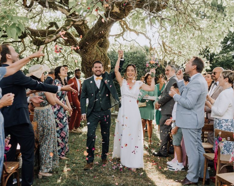 Couple célébrant leur mariage sous un arbre, entourés d'invités heureux lançant des pétales.