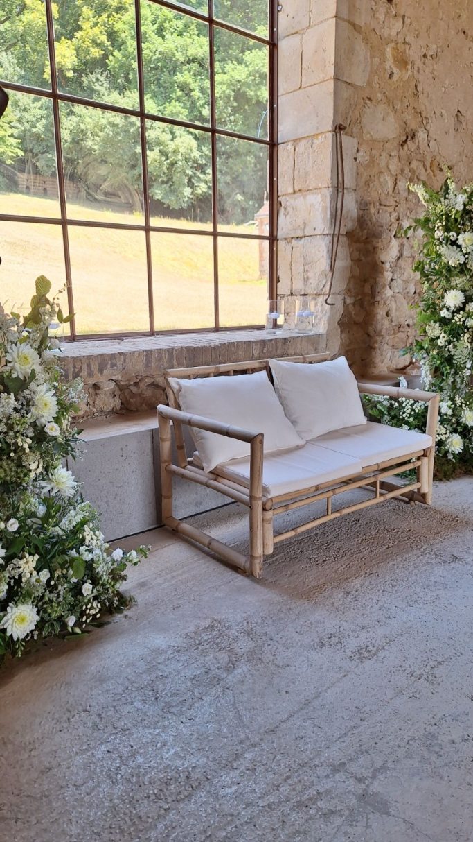 Canapé en bois clair avec coussins, entouré de fleurs blanches dans un espace lumineux.